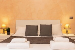 A neatly made bed with white and dark pillows, two bedside lamps, and two folded white towels on a brown mat. - Rental in Rome Trastevere Home Apartment (Rome)