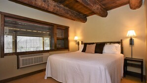 Ein Schlafzimmer mit einem großen Bett, Holzbalken an der Decke, einem Fenster mit Jalousien und zwei Wandleuchten.