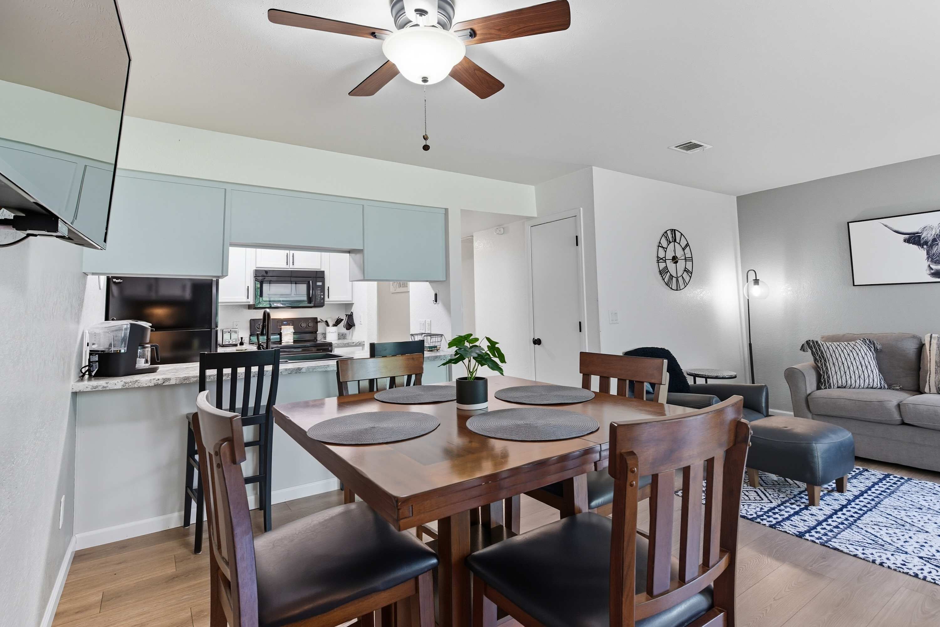 A modern kitchen with light blue cabinets, a dining table with four chairs, and a ceiling fan.