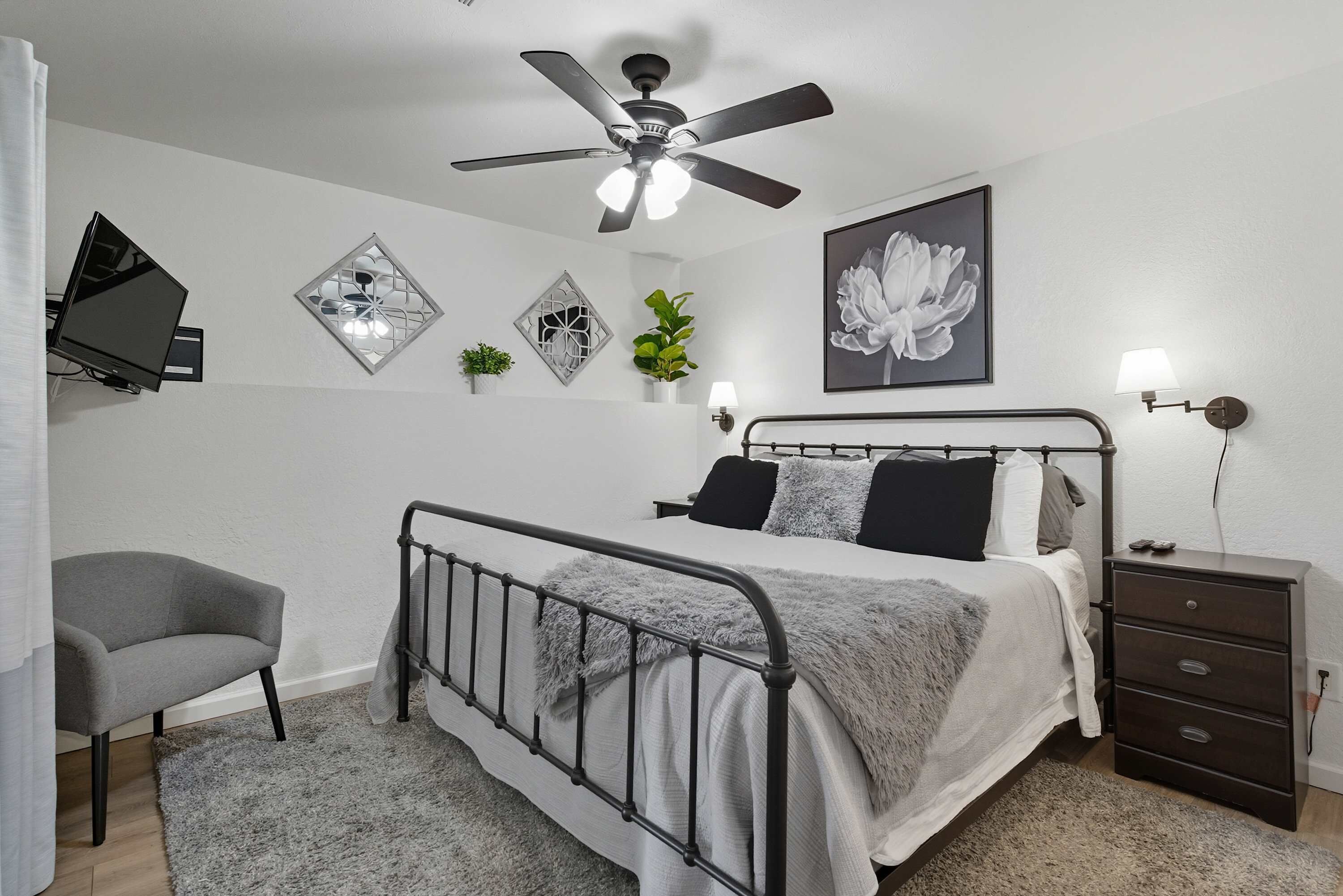Un dormitorio con cama, mesita de noche, ventilador de techo, televisor montado en la pared, decoración en la pared y una pequeña silla.