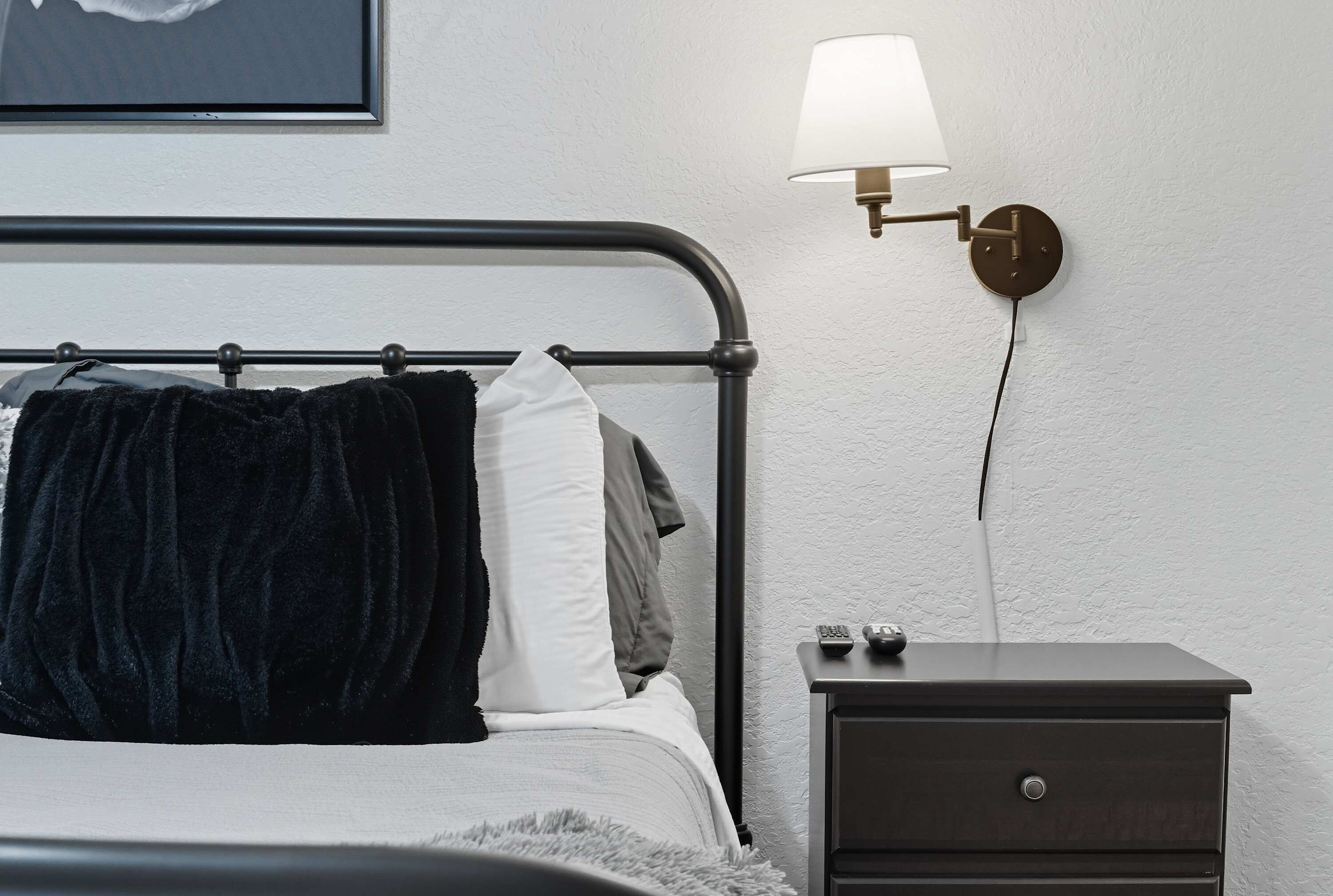 Un lit bien fait, avec une couverture et des oreillers sombres, une table de chevet avec une lampe et un tableau encadré au mur.