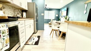 Een moderne keuken met witte kasten, een ingebouwde oven en een koelkast. Er is een eethoek met een tafel en stoelen, en een bord met daarop 'keuken' aan de muur.