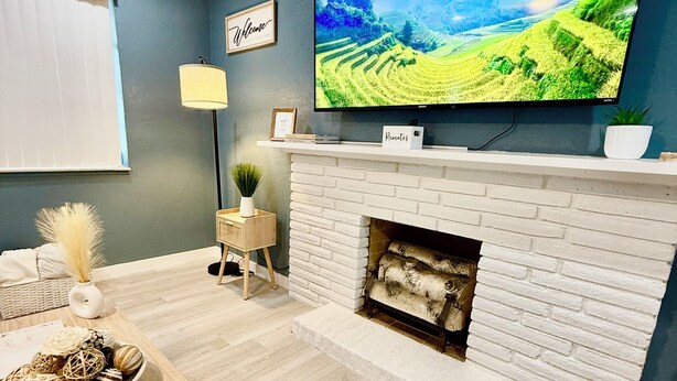 Een moderne woonkamer met een grote flatscreen televisie aan de muur, een wit bakstenen haard en een houten salontafel met decoratieve objecten.