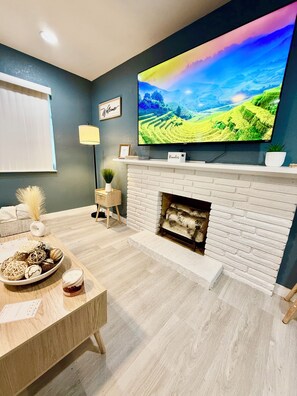 Una sala moderna con un televisor de pantalla plana sobre una chimenea de ladrillo blanco, una mesa de centro de madera con objetos decorativos y una lámpara de pared.
