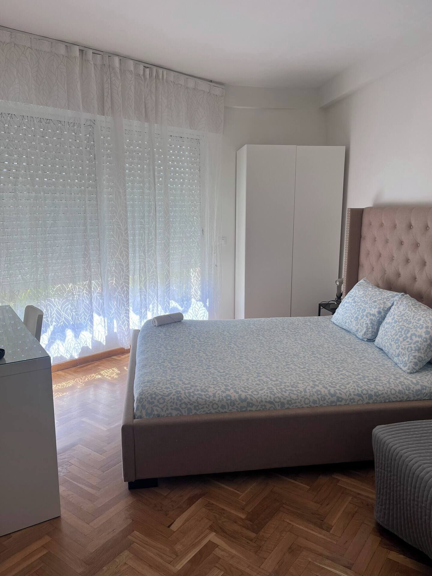 Un dormitorio con cama, armario, escritorio y ventana con cortinas traslúcidas.