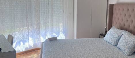 Un dormitorio con cama, armario, escritorio y ventana con cortinas traslúcidas.