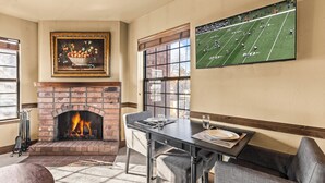 A dining area with a brick fireplace, a table set for two, and a sports game on the wall.