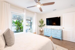 Un dormitorio con cama, ventilador de techo, televisor, cómoda y ventana con plantas.