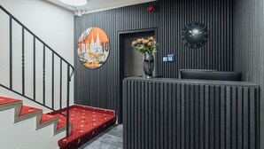 A reception area with a modern design, featuring a reception desk, a clock, a decorative wall with vertical lines, and a staircase with red carpeted steps.