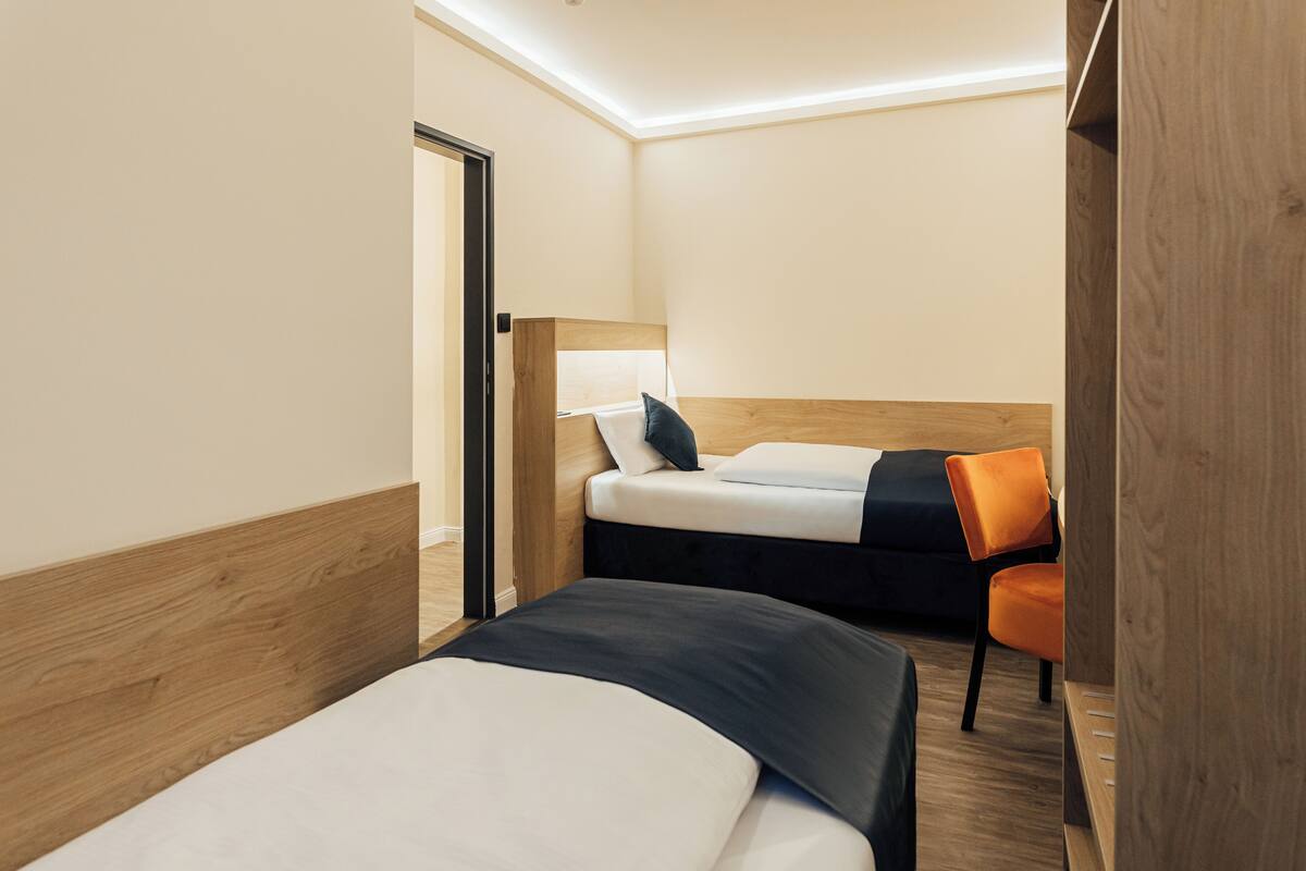 A hotel room with two beds, a wooden headboard, a small orange chair, and a door leading to another room.