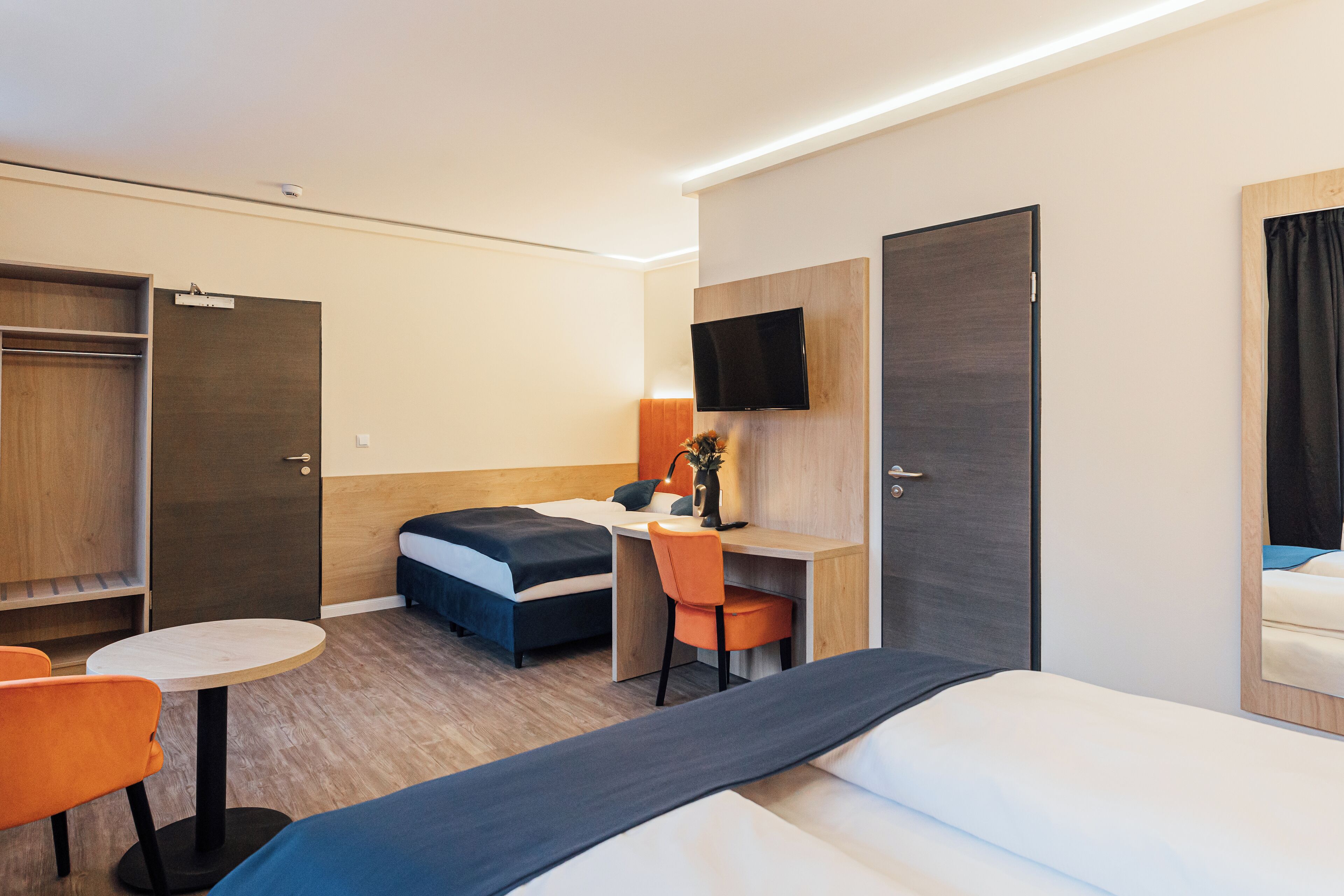A hotel room with a bed, a desk with a chair, a TV mounted on the wall, and a wardrobe.