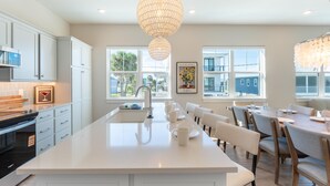 Una cocina moderna con armarios blancos, una isla grande con asientos, iluminación colgante y ventanas amplias que ofrecen vistas al exterior.