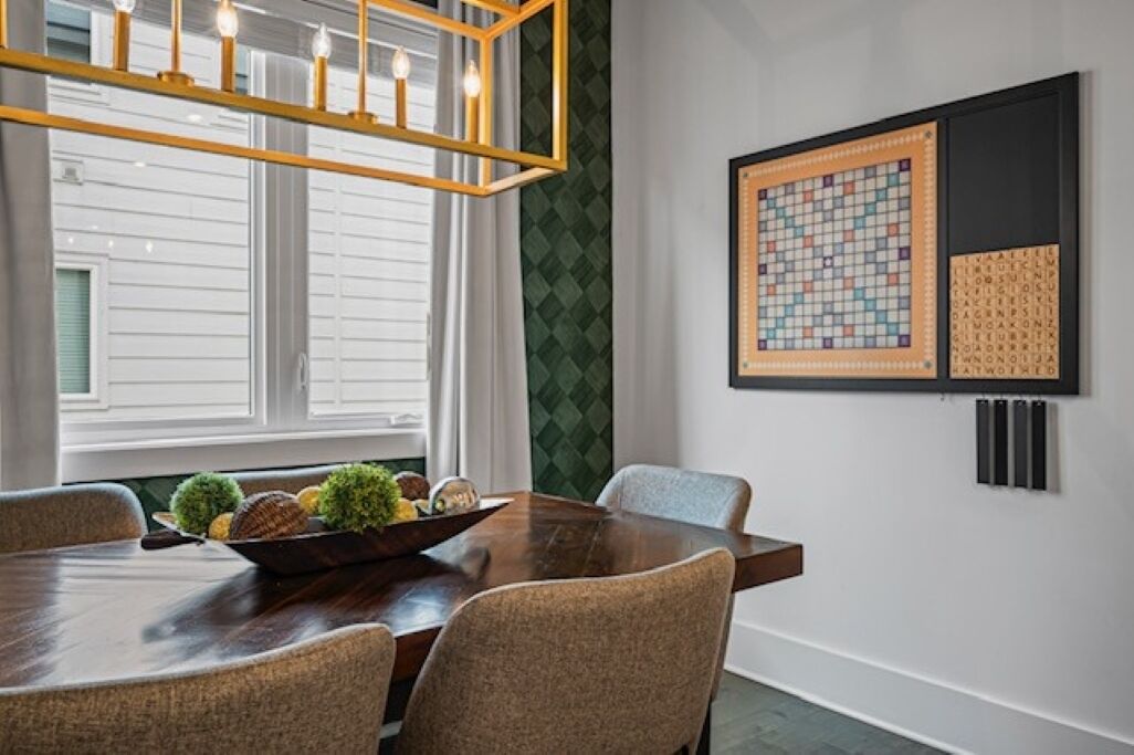 Un coin repas avec une table en bois, des chaises rembourrées, un luminaire décoratif et un mot croisé encadré accroché au mur.