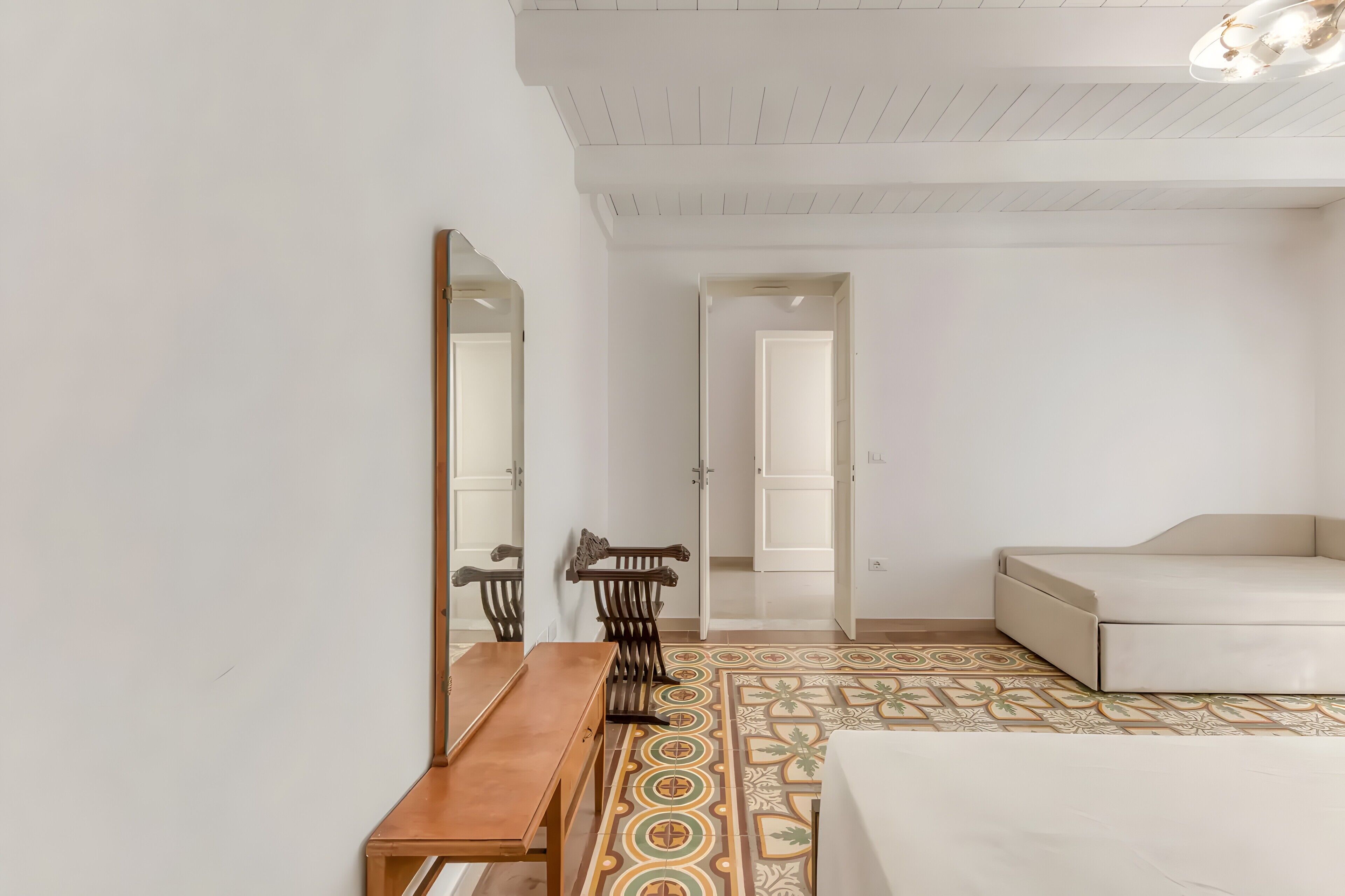 A room with a bed, a wooden table, two chairs, a mirror, and a patterned floor.