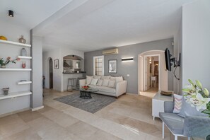 A modern living room with a white sofa, a glass coffee table, and a grey armchair. There is a kitchen in the background with a white countertop and a built-in oven.