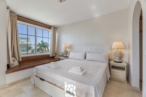 A bedroom with a large bed, two bedside tables with lamps, a window with curtains, and a view of palm trees.