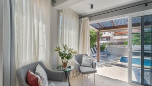 A modern room with a glass table, two grey armchairs, a potted plant, and a view of a swimming pool and outdoor seating area. - Vacation Villa 'La Caleta Seaside  with Pool' with Sea View, Wi-Fi and Air Conditioning (Adeje)