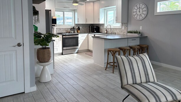 Une cuisine moderne avec des meubles blancs, un four encastré et un îlot central. On y trouve trois tabourets en bois et une chaise à rayures.