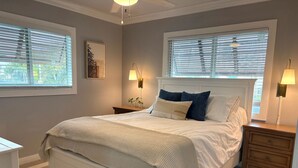 Une chambre à coucher avec un grand lit, deux tables de chevet, une commode, un tableau encadré et deux fenêtres équipées de stores.