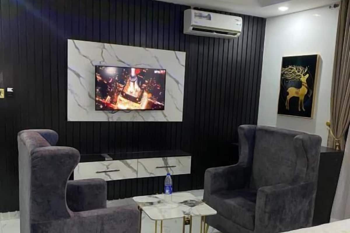 Habitación de hotel moderna con un televisor grande montado en una pared de mármol, dos sillones grises, una mesita de café y una cama con colcha amarilla.
