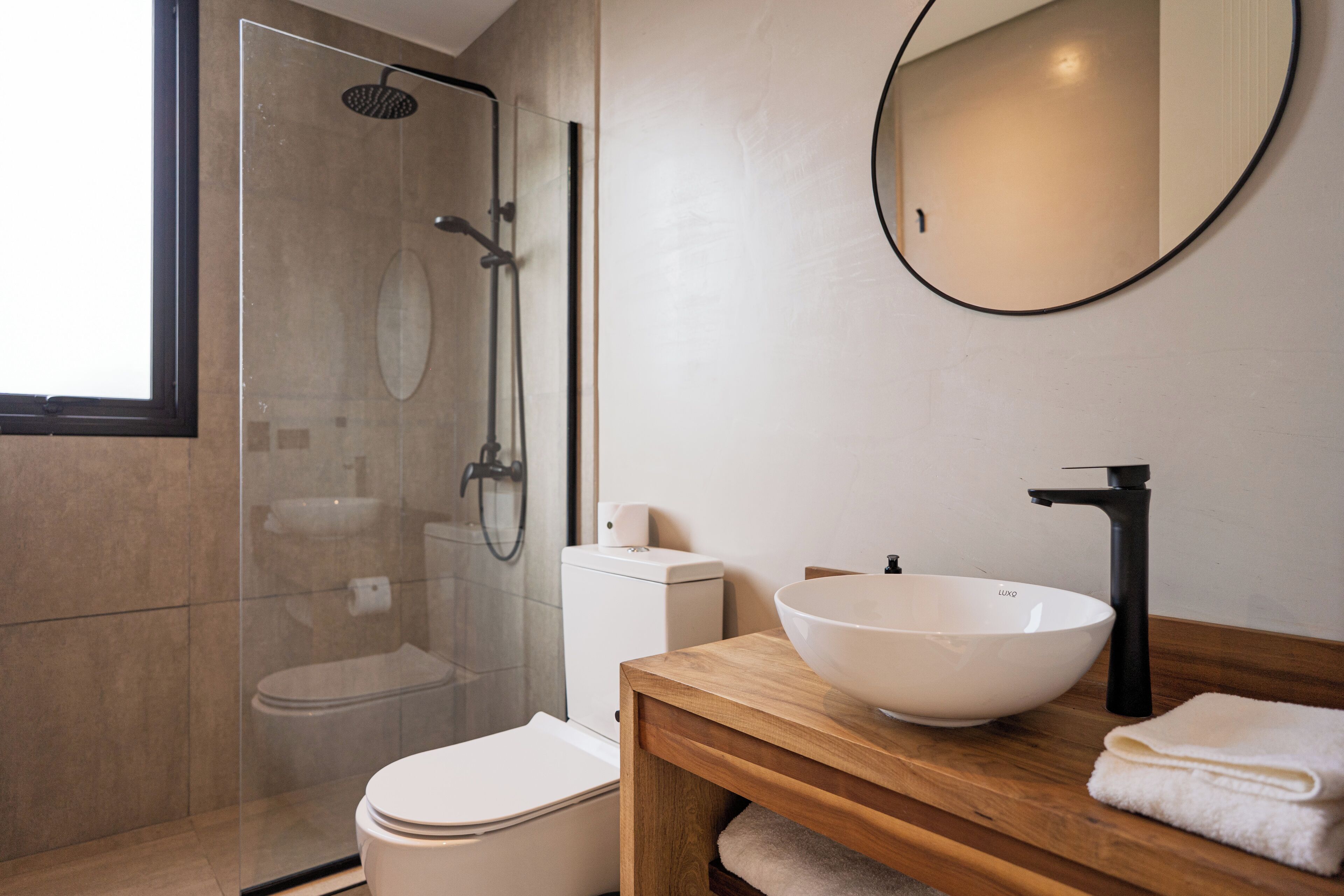 A modern bathroom with a glass-enclosed shower, a white toilet, a wooden vanity with a large white basin, and a round mirror.