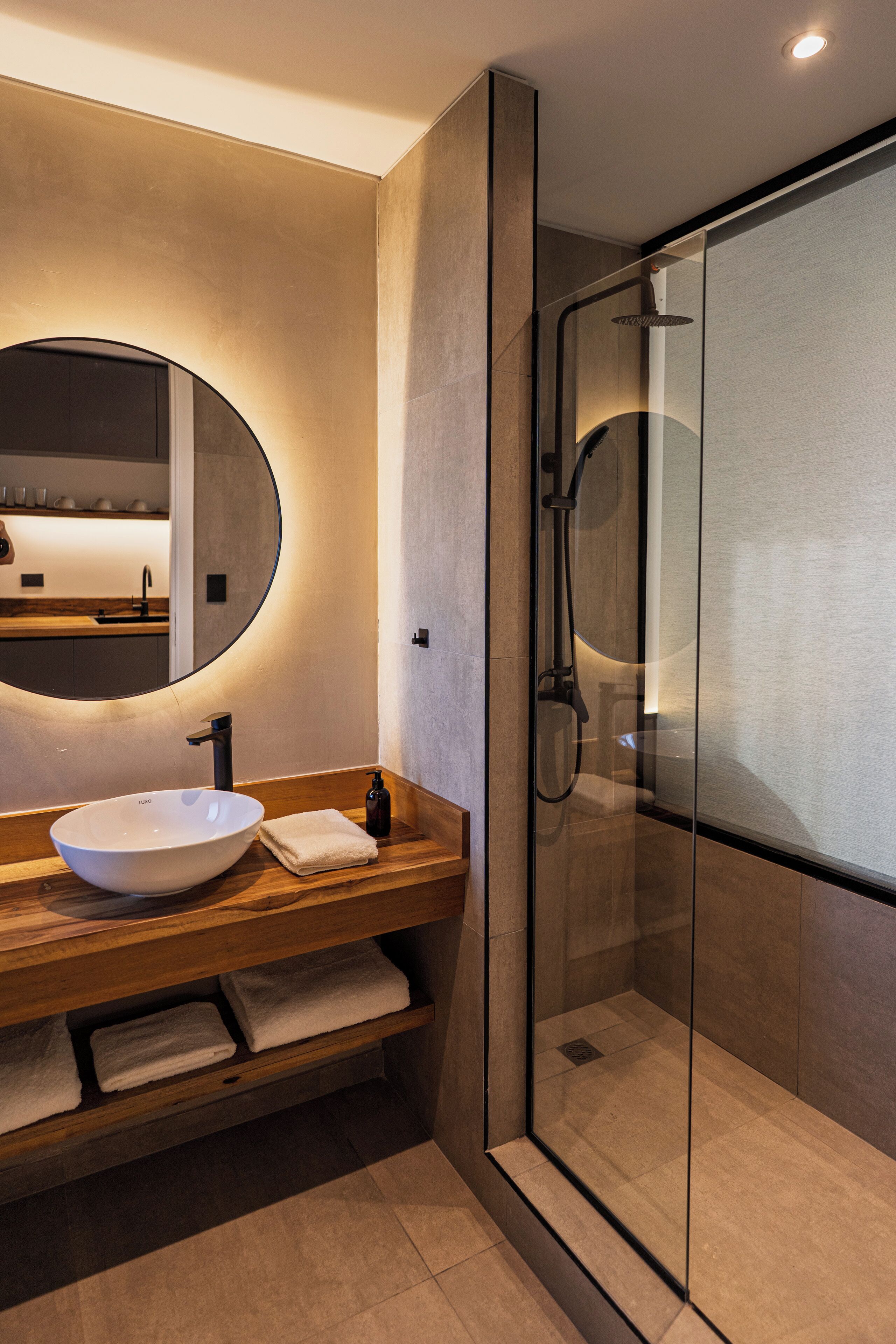 Een moderne badkamer met een grote, ovale spiegel, een houten wastafelmeubel, een witte wasbak en een douchecabine met glazen wanden.