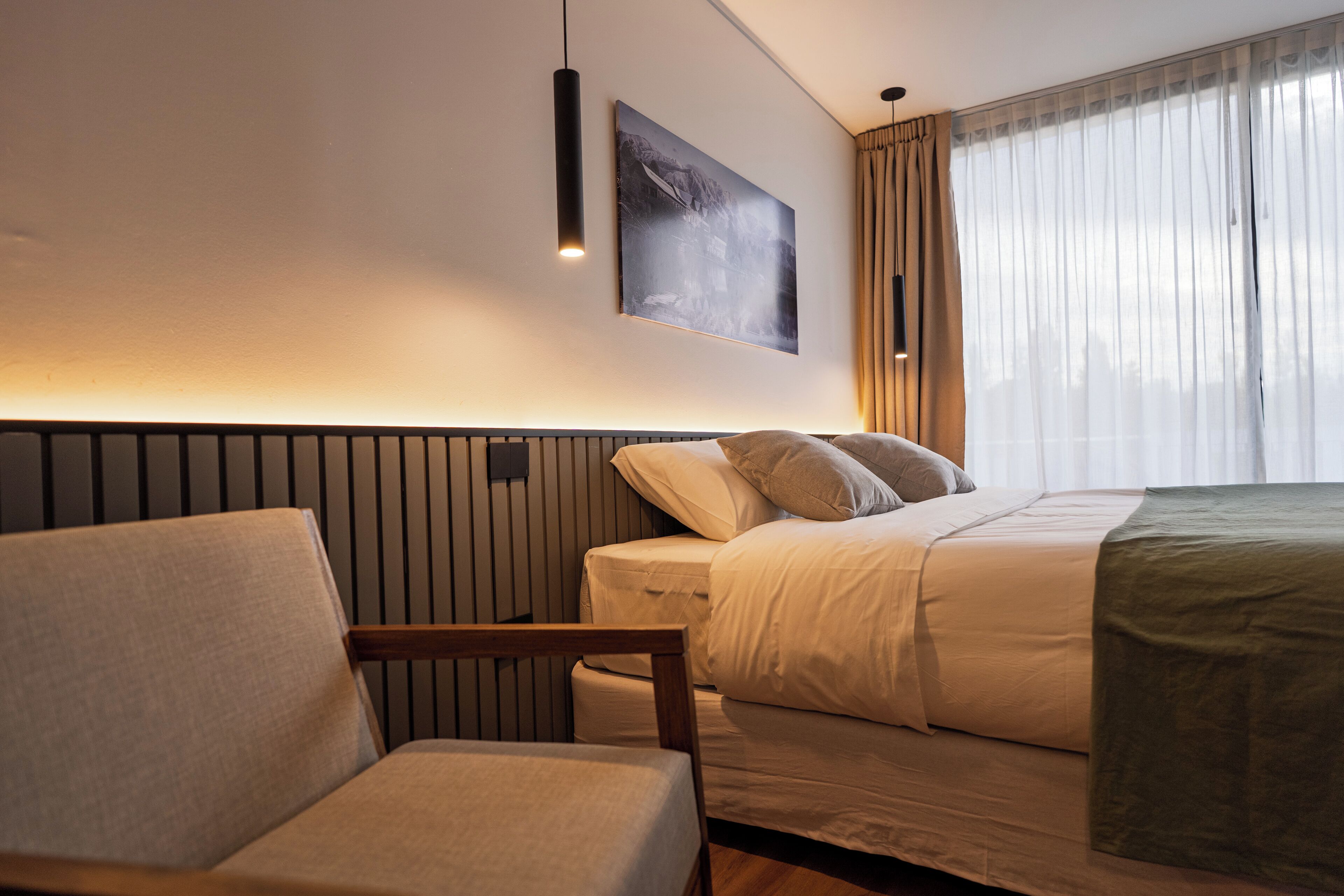 A modern hotel room with a bed, a chair, a wall-mounted artwork, and a window with curtains.