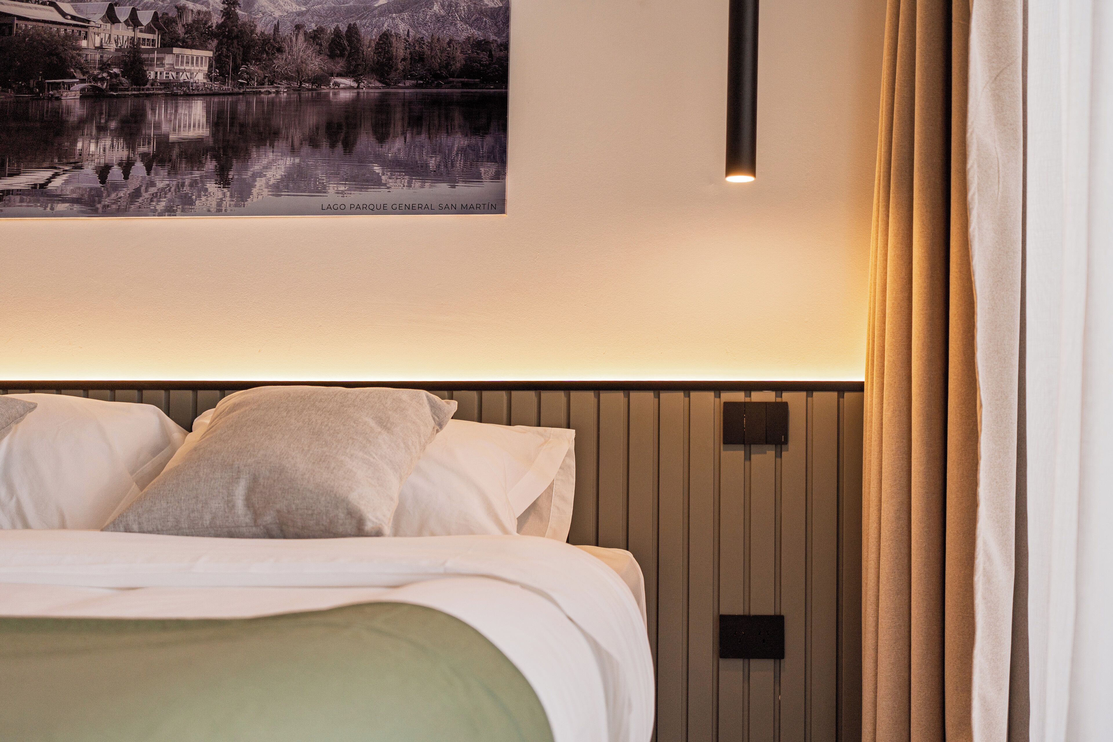 A hotel room with a bed, pillows, a wall-mounted light, and a framed picture of a lake and mountains.