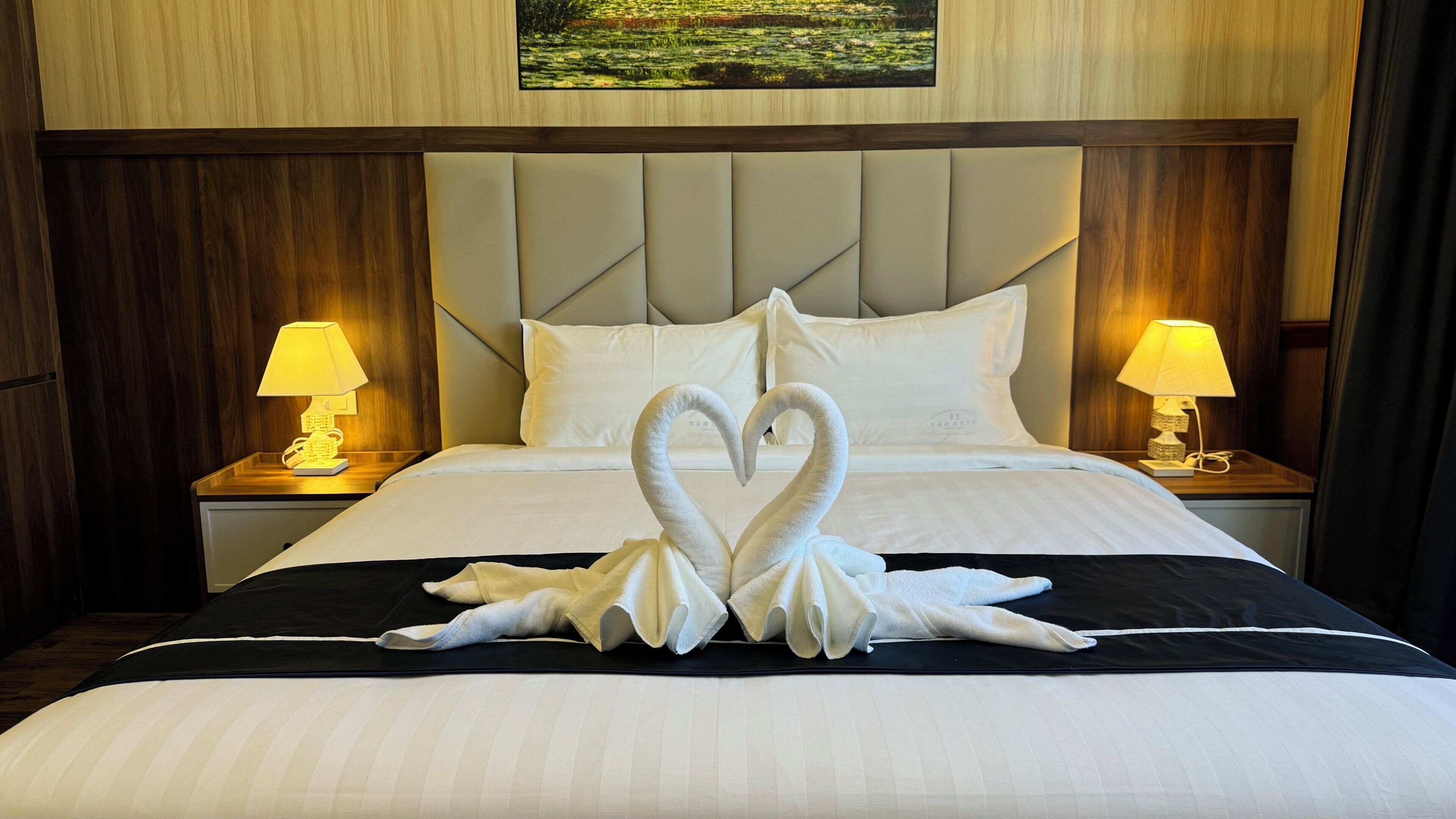 A hotel room with a bed featuring a swan-shaped towel arrangement, wooden headboard, and two bedside lamps.