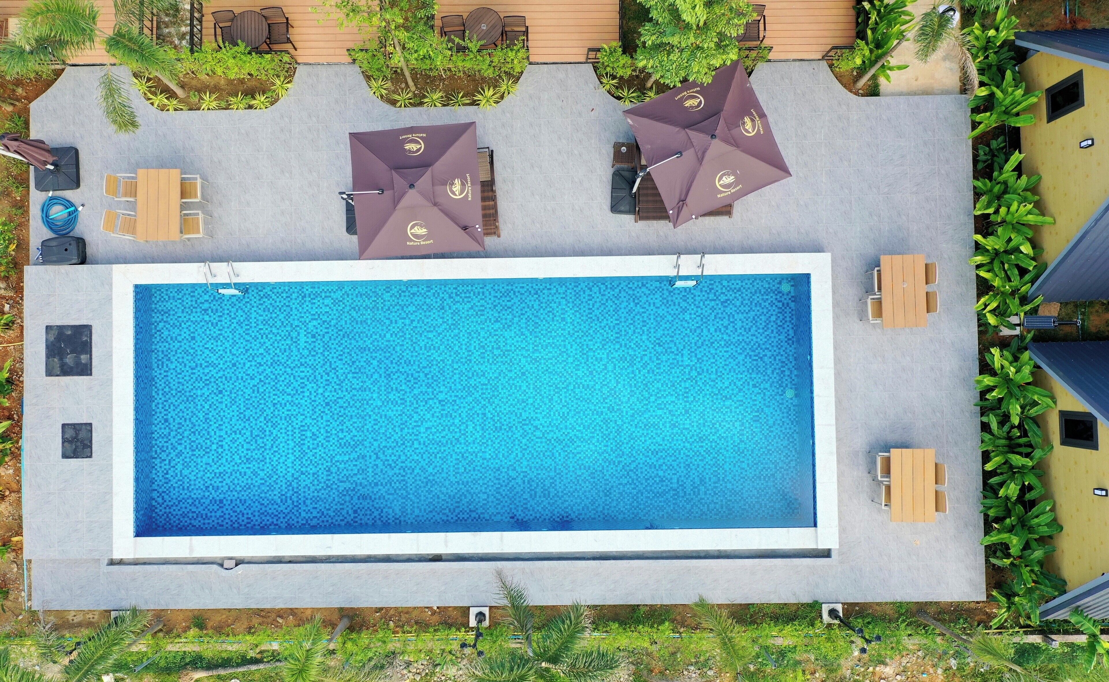 An aerial view of a rectangular swimming pool with a tiled deck surrounding it, equipped with outdoor seating and umbrellas.