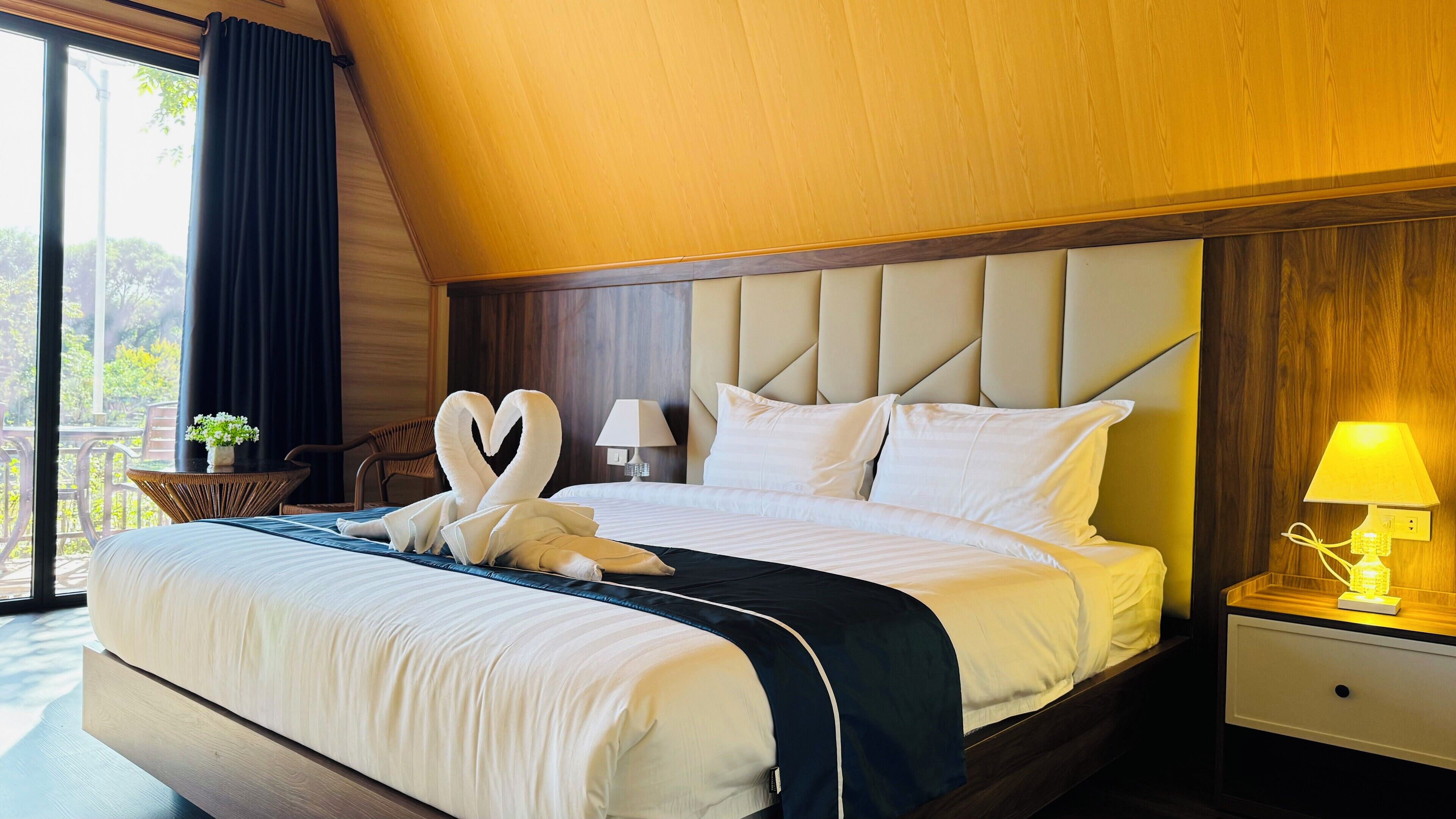 A hotel room with a large bed, a nightstand with a lamp, a view of the outdoors, and a window with curtains.