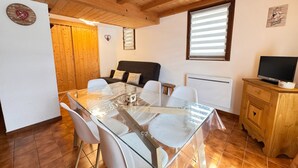 A cozy room with a wooden ceiling, a glass table surrounded by chairs, a sofa with cushions, a television on a wooden cabinet, and a window with blinds. - Alpine stay at the foot of the slopes (Crest-Voland)