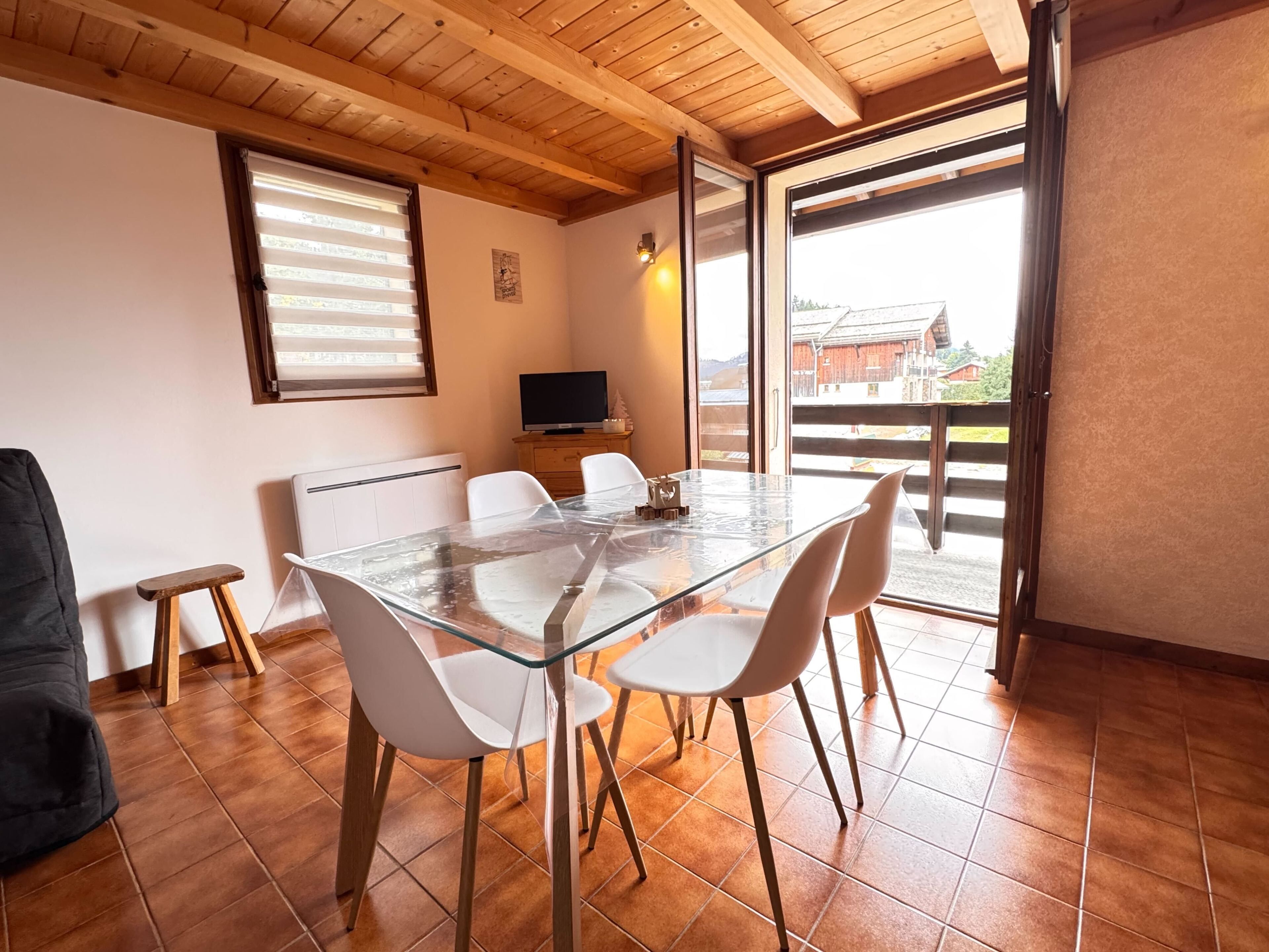 Un coin repas avec une table en verre, des chaises blanches, un téléviseur et une fenêtre avec des stores.