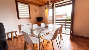 A dining area with a glass table, white chairs, a TV, and a window with blinds. - Alpine stay at the foot of the slopes (Crest-Voland)