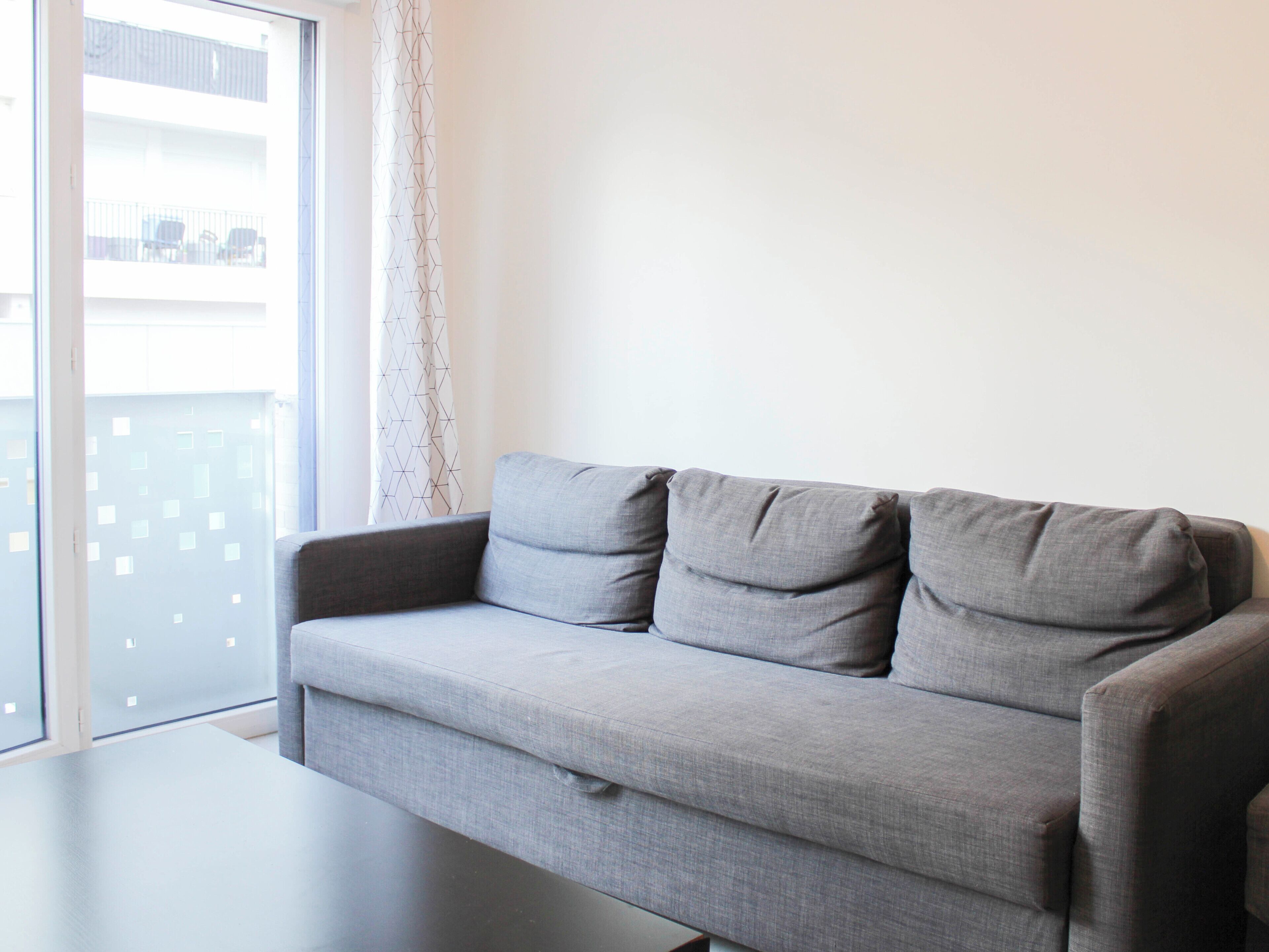 A grey sofa with three cushions in a modern living room with a large window and a view of a balcony.