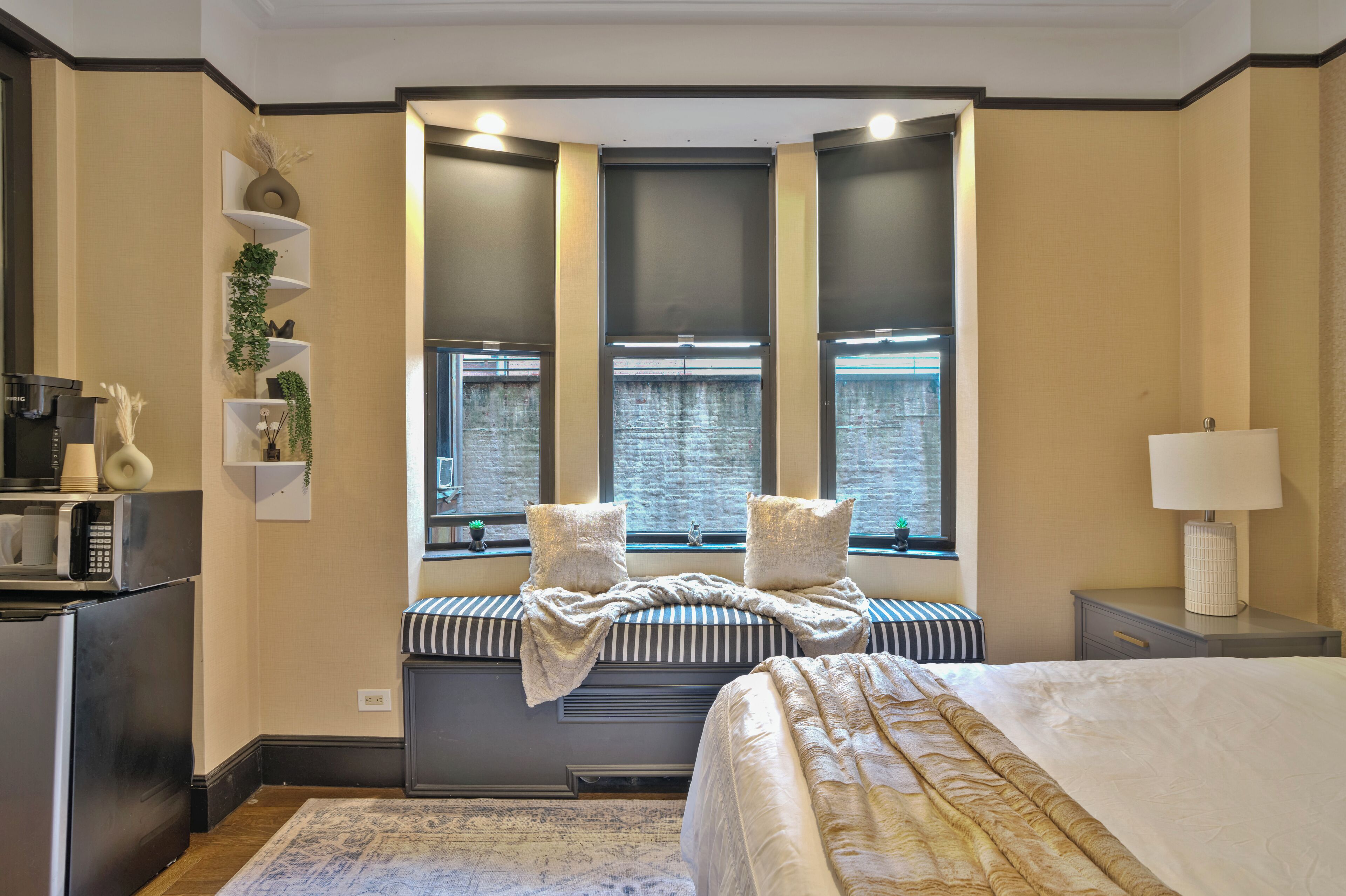 Une chambre avec un grand lit, un coin fenêtre agrémenté de coussins, une table de chevet, une lampe et un coin cuisine équipé d’un four à micro-ondes.