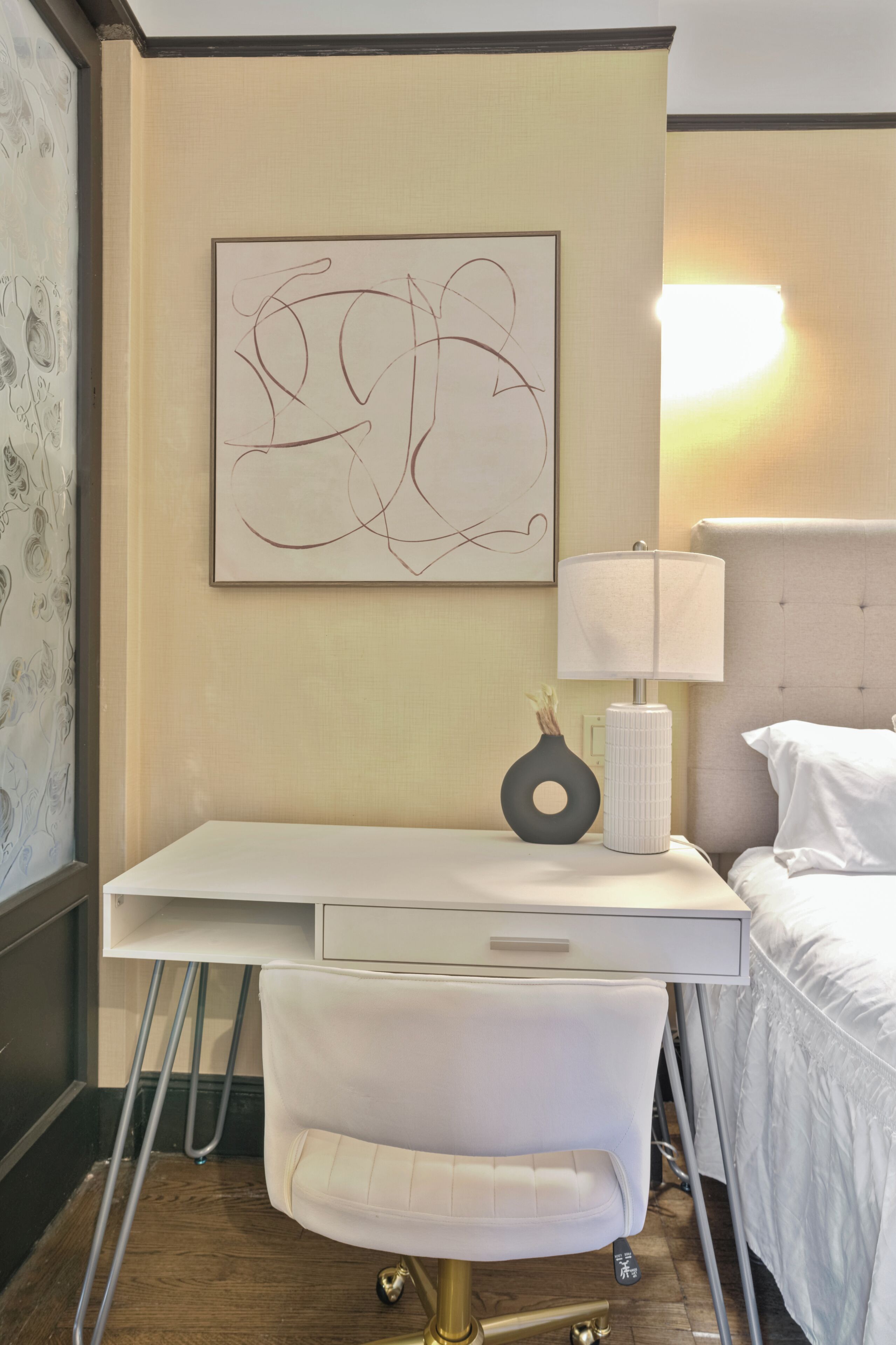 Habitación de hotel con un escritorio blanco, una silla blanca, una cama con ropa de cama blanca, una obra de arte abstracta colgada en la pared y una lámpara de noche.