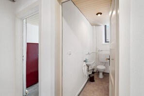 Un couloir étroit mène à une salle de bain équipée de toilettes, d’un lavabo et d’une porte rouge.