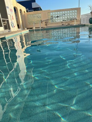 Uma piscina cristalina com um salva-vidas e placas de aviso, cercada por um edifício bege com janelas em arco.