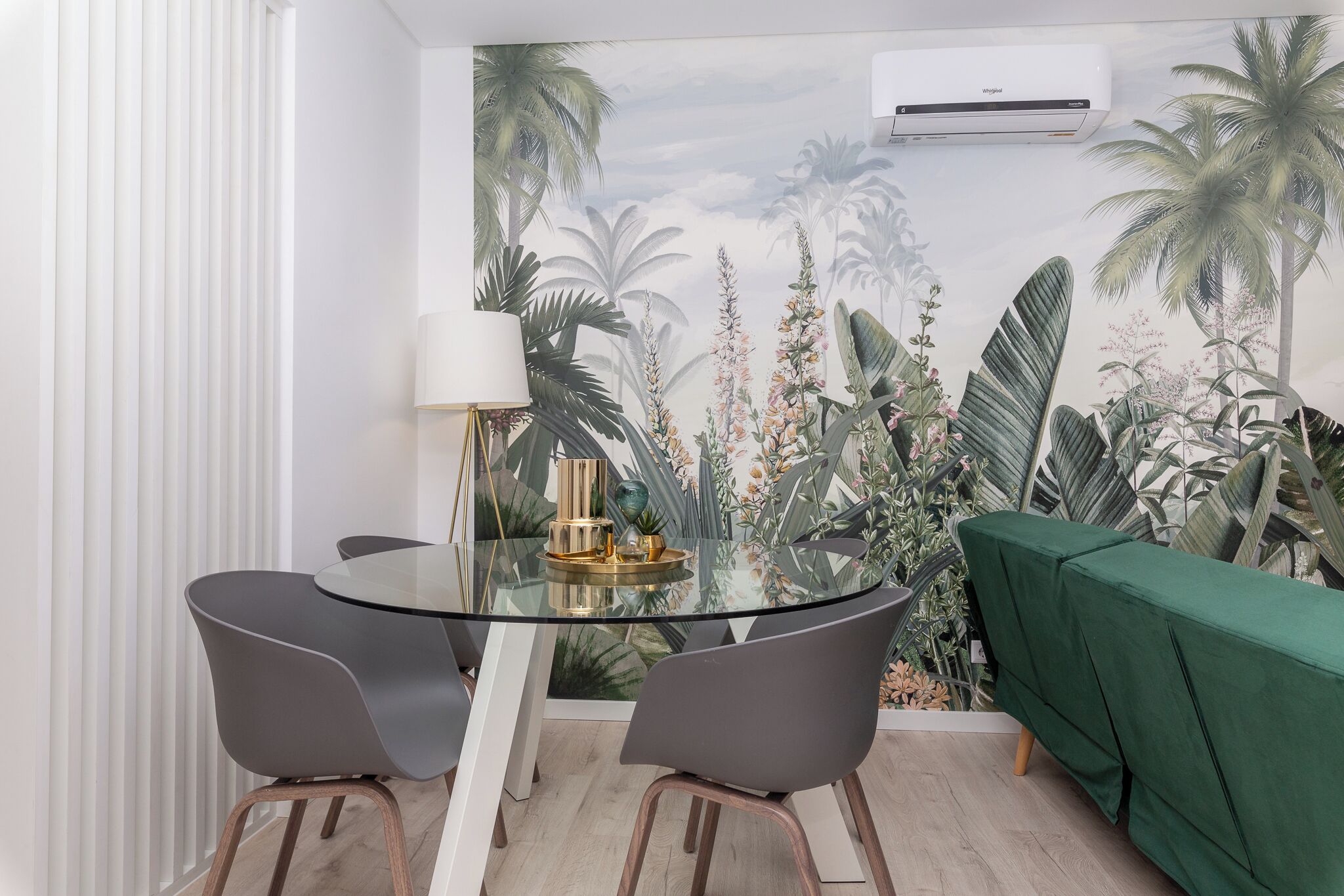 A dining area with a glass table, two grey chairs, a green sofa, and a wall mural of tropical plants.