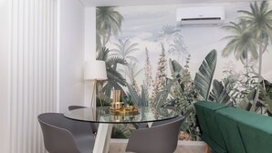 A dining area with a glass table, two grey chairs, a green sofa, and a wall mural of tropical plants. - Margotia Apartment, Faro, Algarve (Faro)