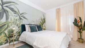 A bedroom with a bed, a nightstand, a lamp, a large plant, and a window with curtains. - Margotia Apartment, Faro, Algarve (Faro)