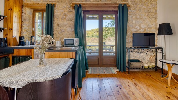 Ein gemütliches Zimmer mit Steinwand, Holzboden, einem Esstisch mit Stühlen, einem Fernseher auf einem Schrank und einem Fenster mit Vorhängen.