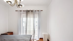 A bedroom with a bed, a wooden nightstand, a laundry basket, and sheer curtains. - Barra Sol - Farol na Praia Apartment with Shared Pool, Balcony, and Wi-Fi (Praia da Barra)