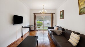 A living room with a sofa, a TV, a table, and a view through a glass door. - Barra Sol - Farol na Praia Apartment with Shared Pool, Balcony, and Wi-Fi (Praia da Barra)