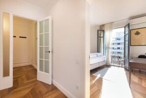 A room with a wooden floor, a white door, a white wall, a window with curtains, a white sofa, and a small table.