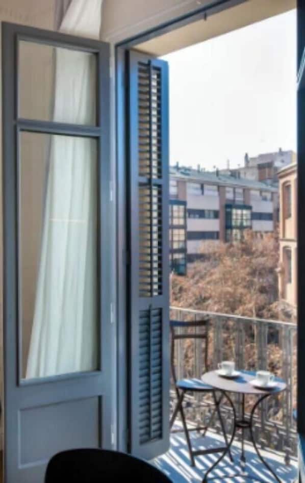 Un balcón con mesa y sillas, una ventana con contraventanas y vistas a edificios y árboles.