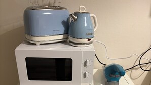Una encimera de cocina blanca con un microondas, un tostador y una tetera. Abajo, hay estantes con frascos y una caja con la etiqueta 'TEA'.