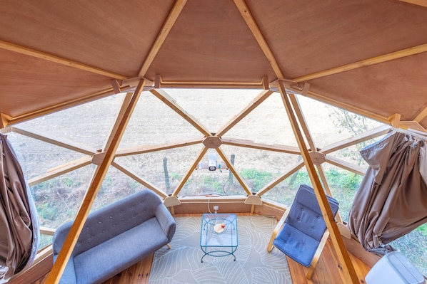 A geodesic dome structure with a wooden floor, two sofas, a small table, and a view through large windows.