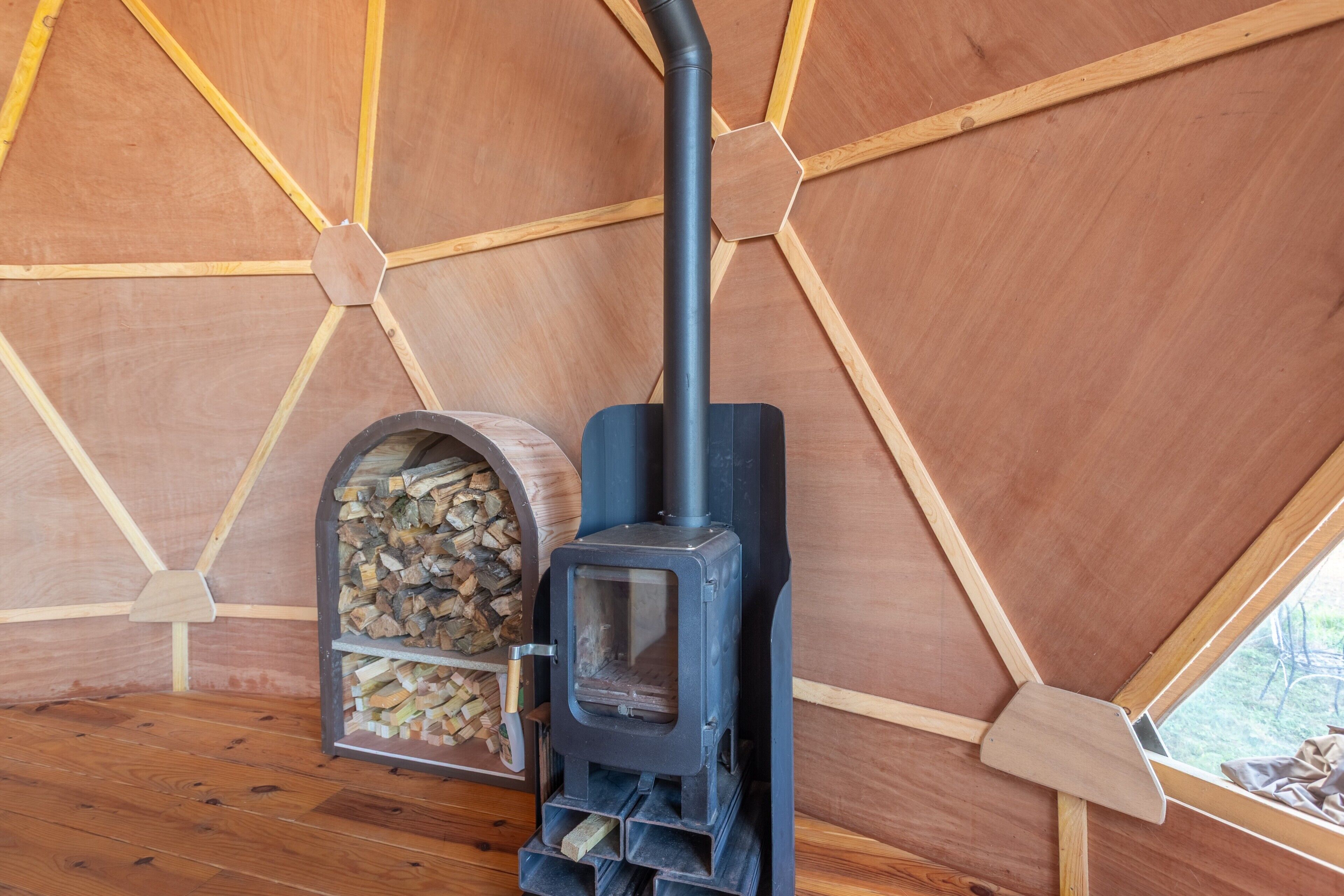Un poêle à bois installé dans une structure ressemblant à une tente, dotée d’un plafond en bois hexagonal et d’une fenêtre.